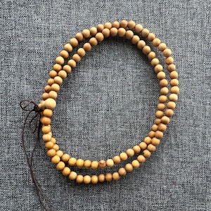 Puede incluir: Un collar de cuentas de madera de doble hebra con acabado natural. Las cuentas son peque&ntilde;as y redondas, conectadas por un cord&oacute;n marr&oacute;n. El collar est&aacute; sobre una superficie gris texturizada.