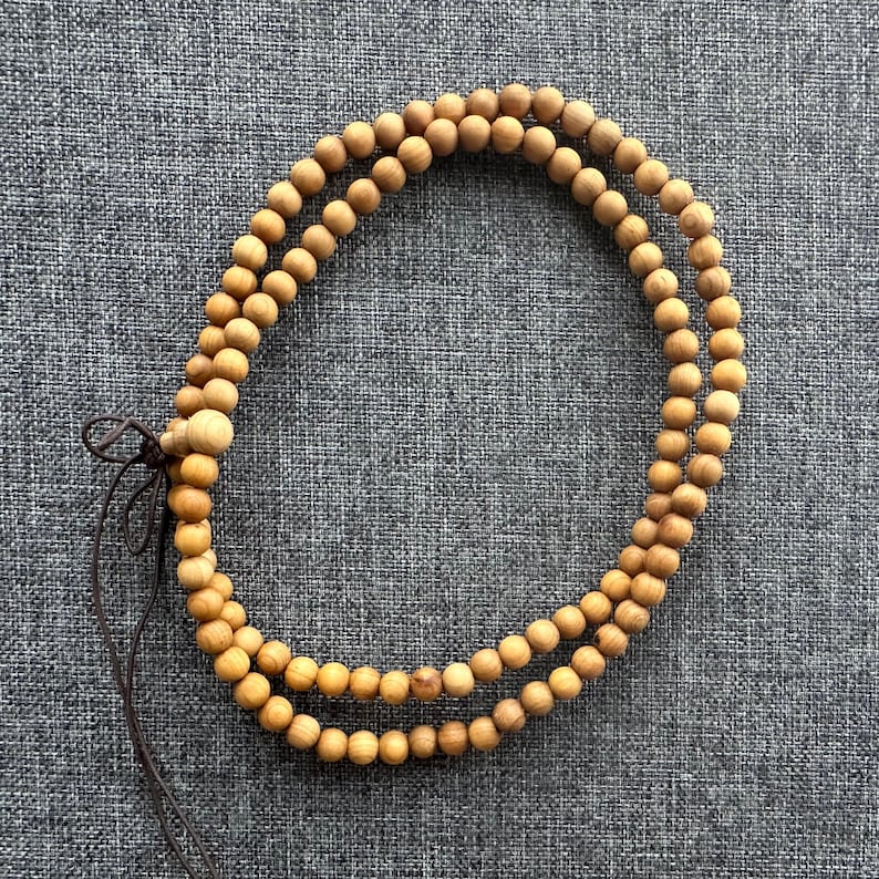 Puede incluir: Un collar de cuentas de madera de doble hebra con acabado natural. Las cuentas son peque&ntilde;as y redondas, conectadas por un cord&oacute;n marr&oacute;n. El collar est&aacute; sobre una superficie gris texturizada.