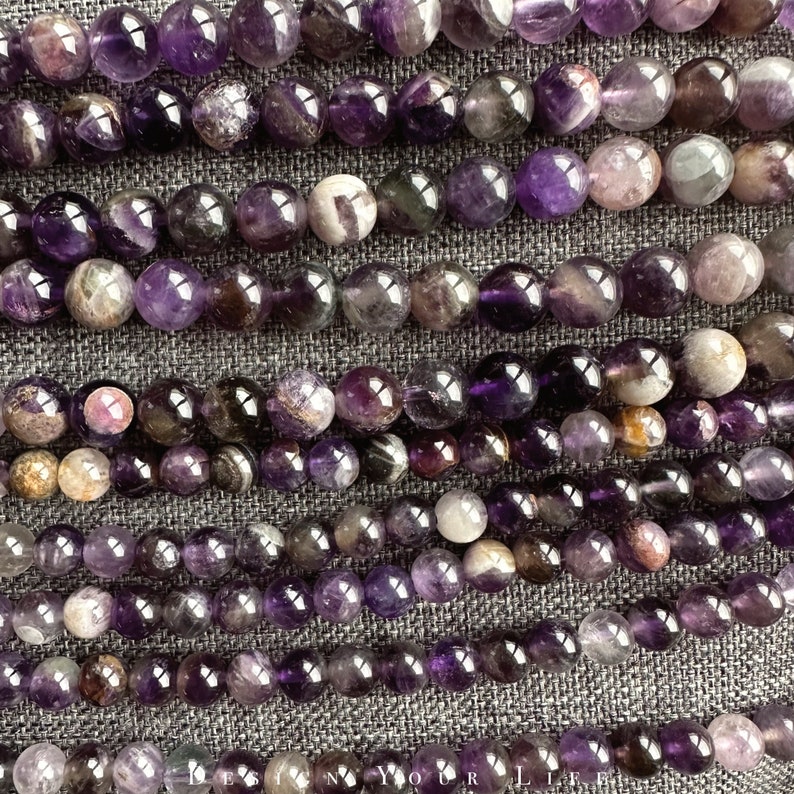 May include: A strand of amethyst beads with a variety of purple hues and patterns. The beads are round and smooth, and they are arranged in a row on a grey fabric background.