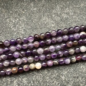 May include: A strand of amethyst beads with a mix of purple and clear tones. The beads are round and smooth, and they are arranged in a loose pile on a grey background.