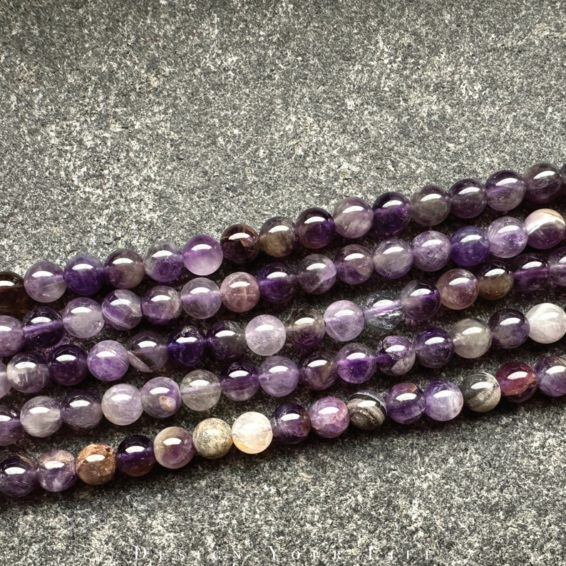 May include: A strand of amethyst beads with a mix of purple and clear tones. The beads are round and smooth, and they are arranged in a loose pile on a grey background.