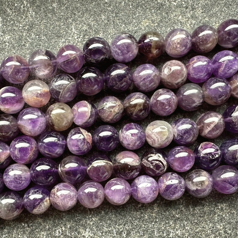 May include: A strand of amethyst beads, each bead is a deep purple colour with white and grey swirls and markings. The beads are round and smooth, and they are arranged in a row.