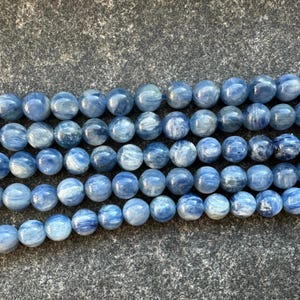 May include: A strand of blue kyanite beads, each bead is round and has a smooth, polished finish. The beads are arranged in a row, with the light reflecting off their surface.
