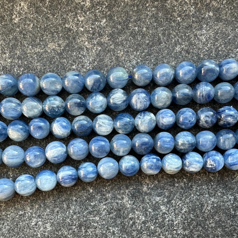 May include: A strand of blue kyanite beads, each bead is round and has a smooth, polished finish. The beads are arranged in a row, with the light reflecting off their surface.