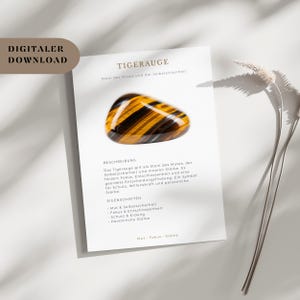 Op de afbeelding: Een gepolijste tijgeroog-edelsteen met een goudbruin en zwart gestreept patroon ligt op een witte kaart. De kaart bevat Duitse tekst, waaronder "TIGERAUGE". De edelsteen is een symbool van moed en innerlijke kracht.
