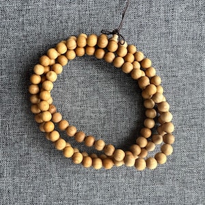 Puede incluir: Un collar de cuentas de madera marr&oacute;n claro, probablemente s&aacute;ndalo, dispuestas en un patr&oacute;n circular. Las cuentas lisas y esf&eacute;ricas est&aacute;n unidas por un cord&oacute;n marr&oacute;n oscuro. El fondo es una tela gris texturizada.