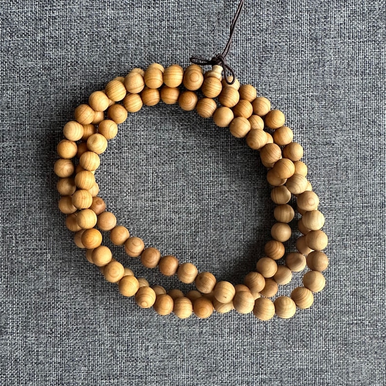 Puede incluir: Un collar de cuentas de madera marr&oacute;n claro, probablemente s&aacute;ndalo, dispuestas en un patr&oacute;n circular. Las cuentas lisas y esf&eacute;ricas est&aacute;n unidas por un cord&oacute;n marr&oacute;n oscuro. El fondo es una tela gris texturizada.