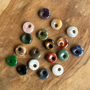 Könnte beinhalten: Sortiment von 18 donutförmigen Edelsteinperlen in verschiedenen Farben, darunter Grün, Blau, Rot, Braun, Schwarz, Weiß und Lila. Jede Perle hat ein Loch in der Mitte.