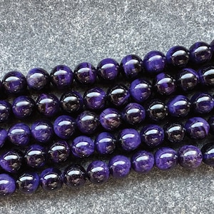 May include: Close-up of strands of polished, round, deep purple beads. The beads have a glossy finish and reflect light, creating highlights. The beads are strung together, suggesting they are for jewelry making or crafting.