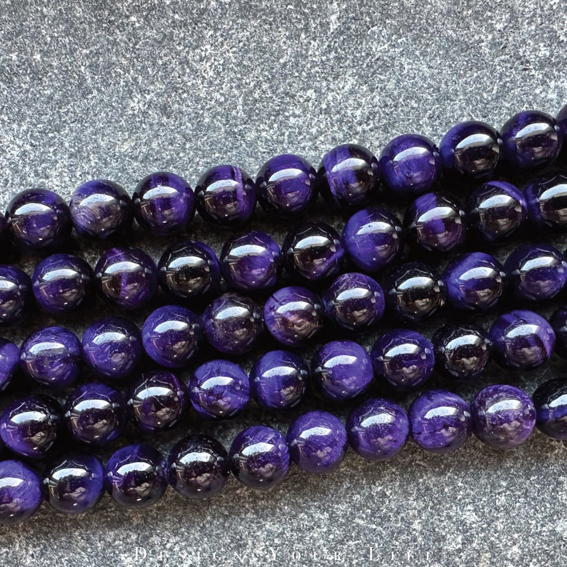 May include: Close-up of strands of polished, round, deep purple beads. The beads have a glossy finish and reflect light, creating highlights. The beads are strung together, suggesting they are for jewelry making or crafting.