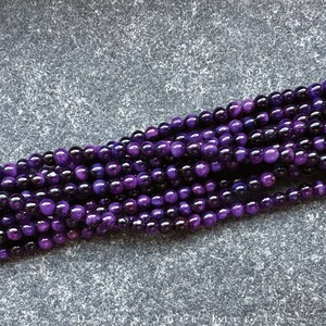 May include: Strands of polished, round beads in varying shades of purple. The beads have a glossy finish and reflect light, creating highlights. The beads are likely for jewelry making or crafting.