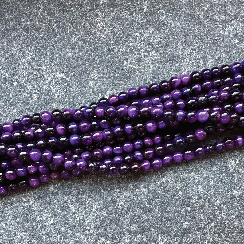May include: Strands of polished, round beads in varying shades of purple. The beads have a glossy finish and reflect light, creating highlights. The beads are likely for jewelry making or crafting.
