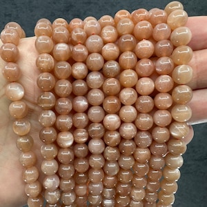 Op de afbeelding: Strengen perzik-kleurige zonnesteen edelsteen kralen. De kralen zijn rond, gepolijst en hebben een subtiele glans. De kralen zijn geregen op een dun, oranje draadje, klaar voor het maken van sieraden.