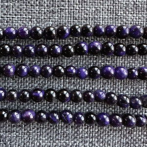 May include: Close-up of several strands of round beads. The beads alternate between deep purple and black, creating a striking contrast. The beads are smooth and reflective, suggesting a polished finish. The arrangement suggests jewelry making or craft supplies.