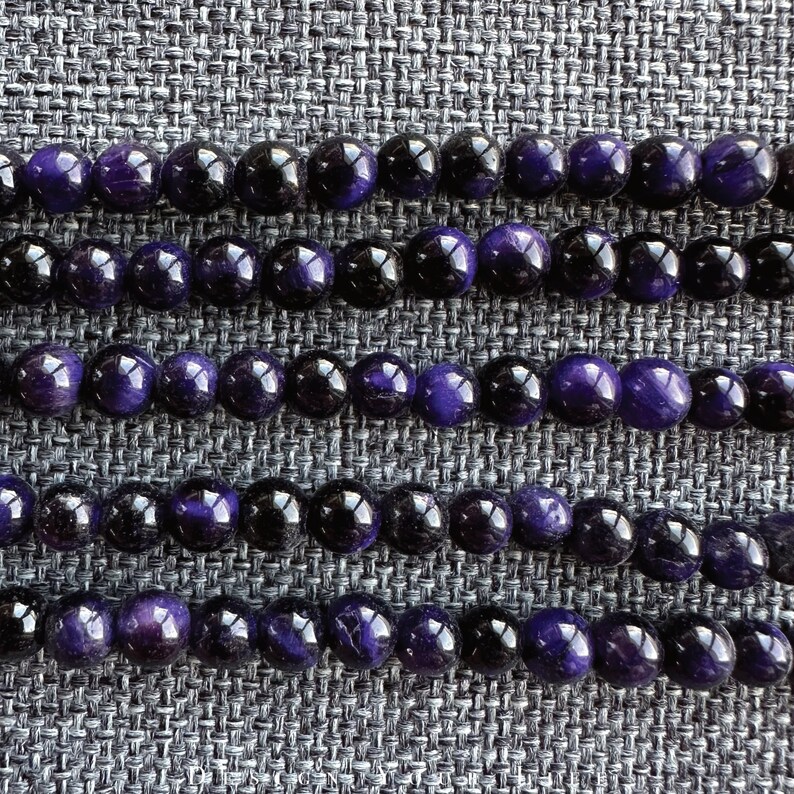 May include: Close-up of several strands of round beads. The beads alternate between deep purple and black, creating a striking contrast. The beads are smooth and reflective, suggesting a polished finish. The arrangement suggests jewelry making or craft supplies.