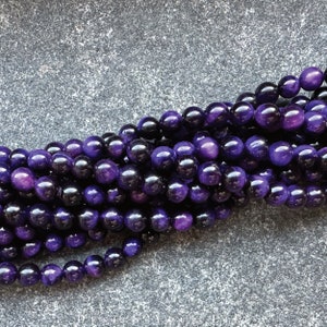 May include: A close-up of a strand of polished, round beads in shades of deep purple and black. The beads have a glossy finish and are strung together, creating a textured, organic pattern. The beads are likely used for jewelry making.