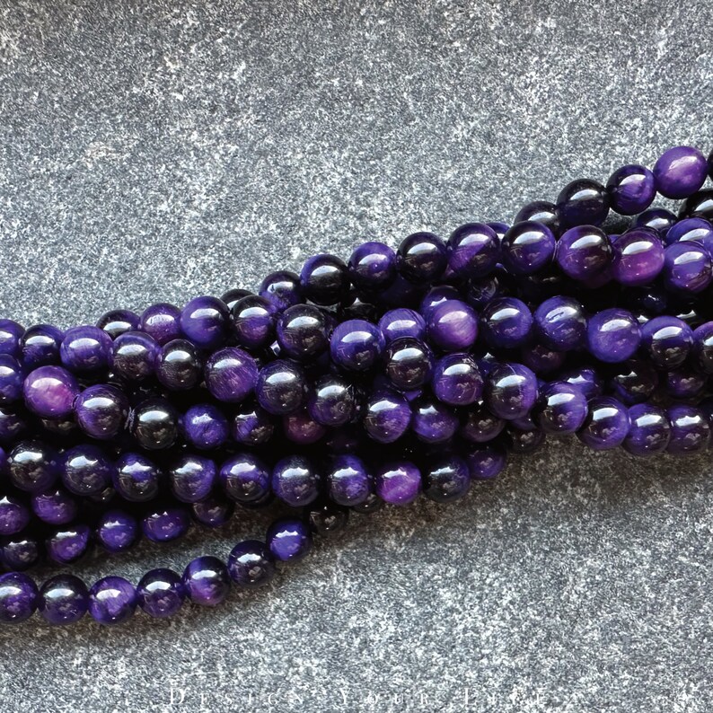 May include: A close-up of a strand of polished, round beads in shades of deep purple and black. The beads have a glossy finish and are strung together, creating a textured, organic pattern. The beads are likely used for jewelry making.