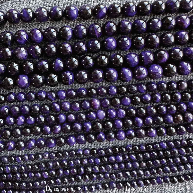 May include: Rows of polished beads in varying sizes and shades of purple and black. The beads are arranged in parallel lines, creating a textured surface. The image is well-lit, highlighting the glossy finish of the beads. The word "DESIGN" is visible at the bottom.