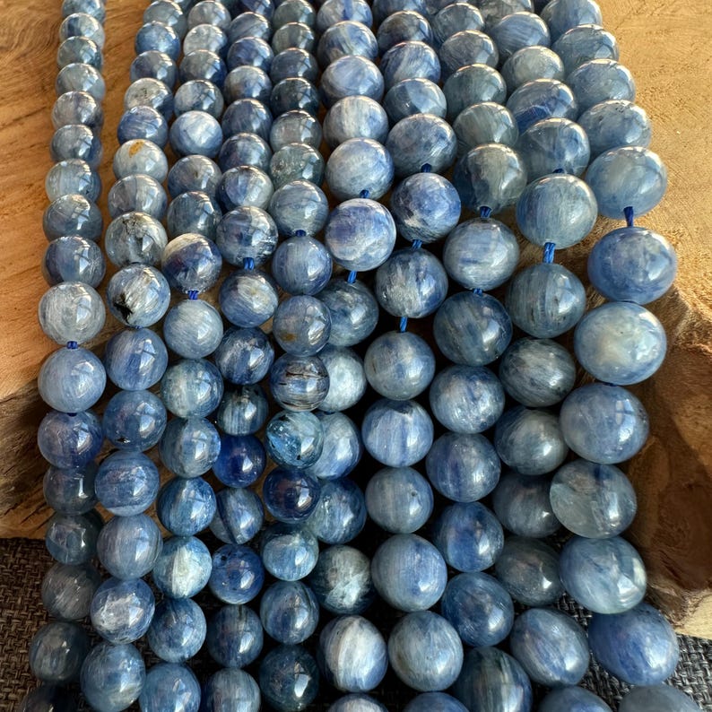 May include: A strand of blue kyanite beads, each bead is round and has a striated pattern. The beads are arranged in rows, creating a visual texture.