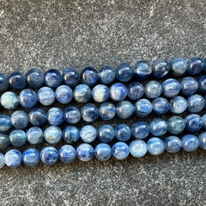 May include: A strand of blue kyanite beads, each bead is round and smooth. The beads are arranged in a row, with the top row slightly overlapping the bottom row.