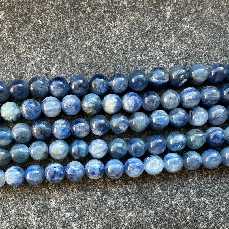 May include: A strand of blue kyanite beads, each bead is round and smooth. The beads are arranged in a row, with the top row slightly overlapping the bottom row.