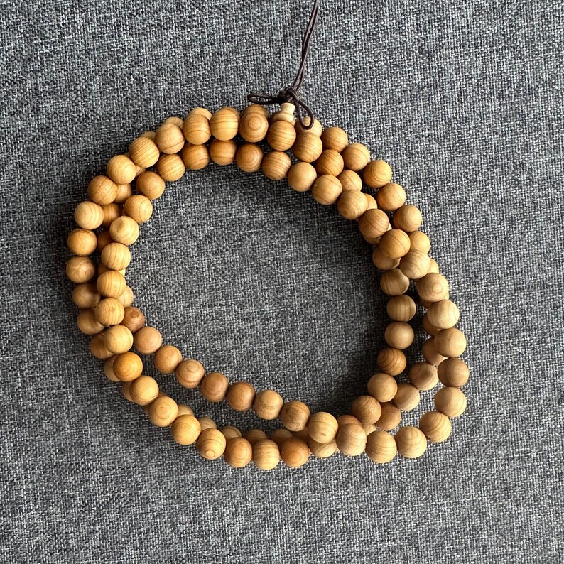 Puede incluir: Un collar de cuentas de madera marr&oacute;n claro, probablemente para meditaci&oacute;n o oraci&oacute;n. Las cuentas son redondas y est&aacute;n ensartadas, formando una forma circular. Una cuerda marr&oacute;n es visible en la parte superior, para colgar o usar.