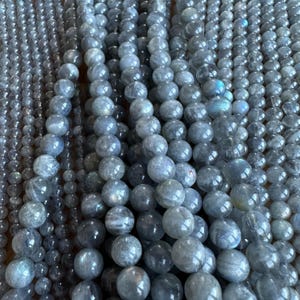 May include: A close-up of multiple strands of round, grey labradorite gemstone beads. The beads are smooth and polished, with a slight iridescent sheen.