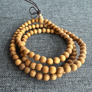 Puede incluir: Un collar de cuentas de oraci&oacute;n de madera con tres hilos de cuentas redondas de color marr&oacute;n claro. Las cuentas est&aacute;n ensartadas con un cord&oacute;n marr&oacute;n, y el collar est&aacute; dispuesto en un patr&oacute;n circular.