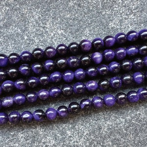 May include: Strands of polished, round beads in shades of deep purple and black. The beads have a glossy finish, reflecting light. The image shows multiple strands, likely for jewelry making or crafting. The beads are uniform in size and shape.