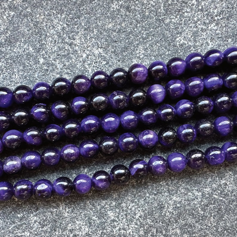 May include: Strands of polished, round beads in shades of deep purple and black. The beads have a glossy finish, reflecting light. The image shows multiple strands, likely for jewelry making or crafting. The beads are uniform in size and shape.