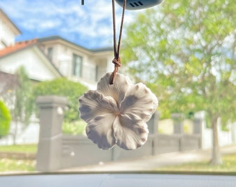 Puede incluir: Un ambientador de aire de cerámica blanca con forma de flor de hibisco que cuelga del espejo retrovisor de un coche. La flor está colgada de un cordón de cuero marrón.