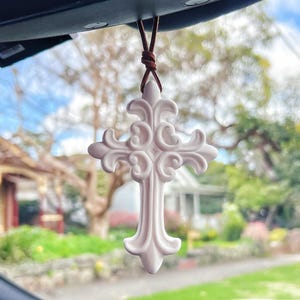 May include: A white, decorative cross-shaped car air freshener suspended by a brown leather cord. The cross has an intricate, swirling pattern. The background shows a street with houses and trees, suggesting an outdoor setting.