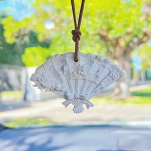 May include: White ceramic fan-shaped car air freshener hanging from a brown string in a car. The air freshener is hanging from the rearview mirror.