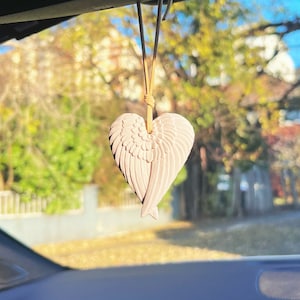 May include: A pink ceramic angel wing heart shaped car air freshener hanging from the rearview mirror of a car.