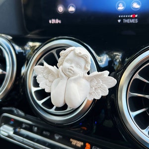 May include: White ceramic angel figurine with wings, sitting in a car air vent. The angel has its head resting on its hands and is looking down.