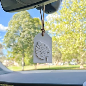 May include: White car air freshener hanging from the rear view mirror. The air freshener is shaped like a tag and features a white, embossed tropical leaf design.
