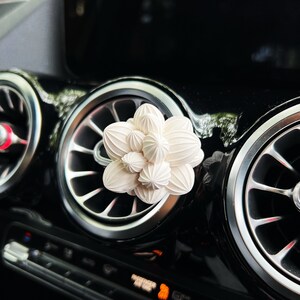 May include: A white car air freshener shaped like a flower with multiple petals. The air freshener is placed in the air vent of a car.