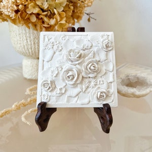 May include: A white, square relief sculpture with detailed roses and floral elements. The artwork is displayed on a dark brown wooden easel. The sculpture is approximately 15 cm square.