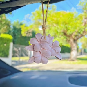 May include: A pink car air freshener shaped like a cluster of plumeria flowers hanging from a rearview mirror.