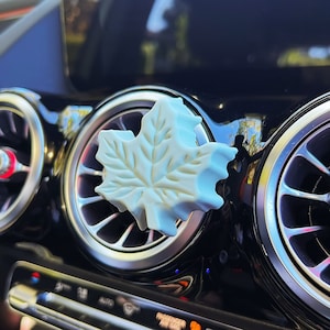 May include: A white maple leaf air freshener hanging from the air vent of a car's dashboard.
