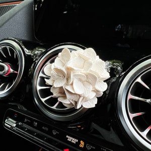 May include: White car air freshener shaped like a hydrangea flower. The air freshener is hanging from the rear-view mirror of a car.