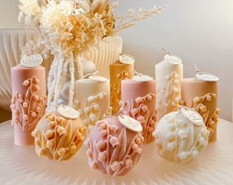 May include: Assortment of decorative candles with a lily of the valley design. The candles are in various shades of pink, cream, and beige. Some candles are round and others are cylindrical. The candles are arranged on a white surface with dried flowers in the background.
