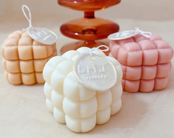 Pode incluir: Três velas decorativas em forma de cubo com um design acolchoado. As velas são em tons de rosa, bege e branco. A vela branca tem uma etiqueta prateada com o texto "LMJ" nela.