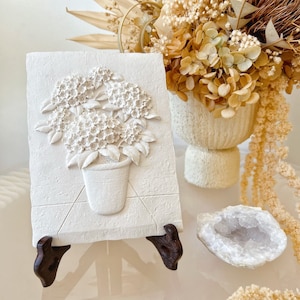 May include: White ceramic wall decor featuring a 3D relief sculpture of a potted plant with white flowers. The decor is displayed on a wooden stand.