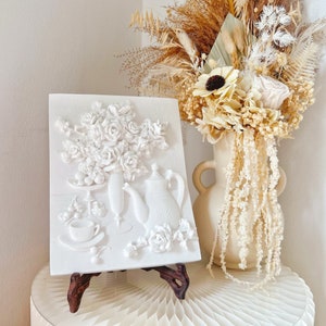 May include: White ceramic wall art featuring a 3D relief sculpture of a bouquet of roses, a teapot, and a teacup on a tray. The art is displayed on a small easel.
