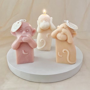 May include: Three small monkey candles, each with a different hand gesture covering their eyes, mouth, and ears. The candles are made of a light beige and pink wax and are sitting on a white circular base.