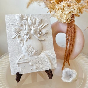 May include: White ceramic wall art featuring a 3D relief sculpture of a vase filled with tulips. The vase is decorated with roses and the background is a white lace pattern.