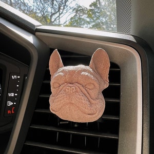May include: A light brown, French bulldog car air freshener hanging from a car's air vent.