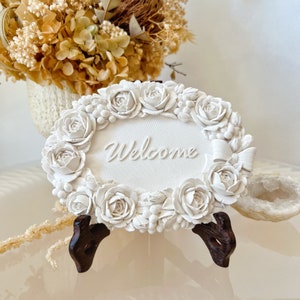 May include: White ceramic welcome sign with a floral design and the word "Welcome" in the centre. The sign is on a small wooden stand.