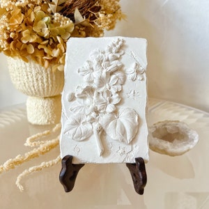 May include: White plaster wall decor featuring a raised relief of a flower with leaves. The decor is displayed on a small wooden stand.